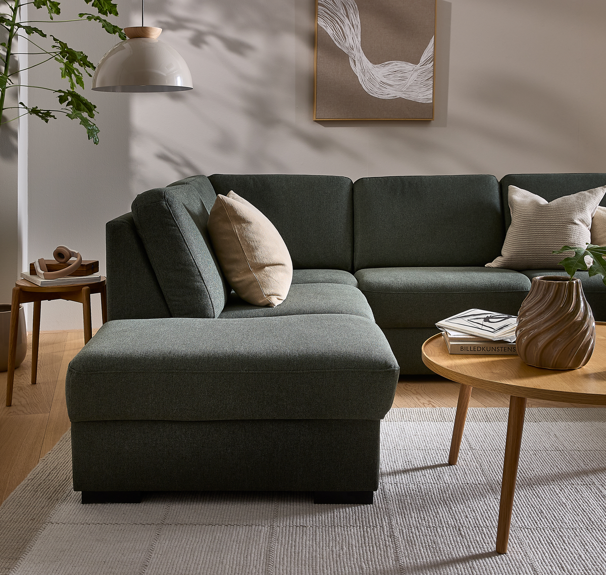 Mørkegrøn hjørnesofa med beige puder i stue; træbord med skulpturel vase og bøgerne ”Billedkunstens” i blødt dagslys, skandinavisk stil.