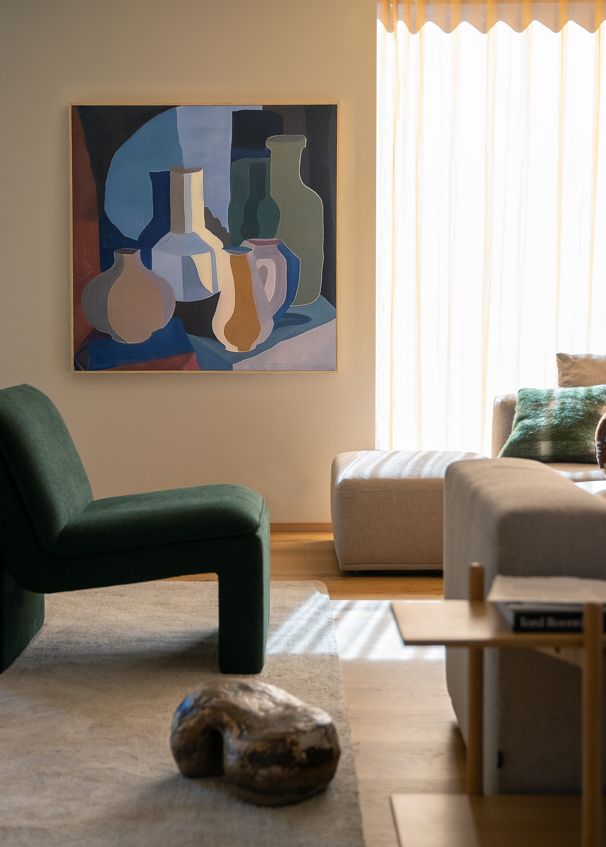 Lyst stue med grøn lænestol, beige sofa, skammel og abstrakt vægkunst; blødt dagslys filtreres gennem lette gardiner.