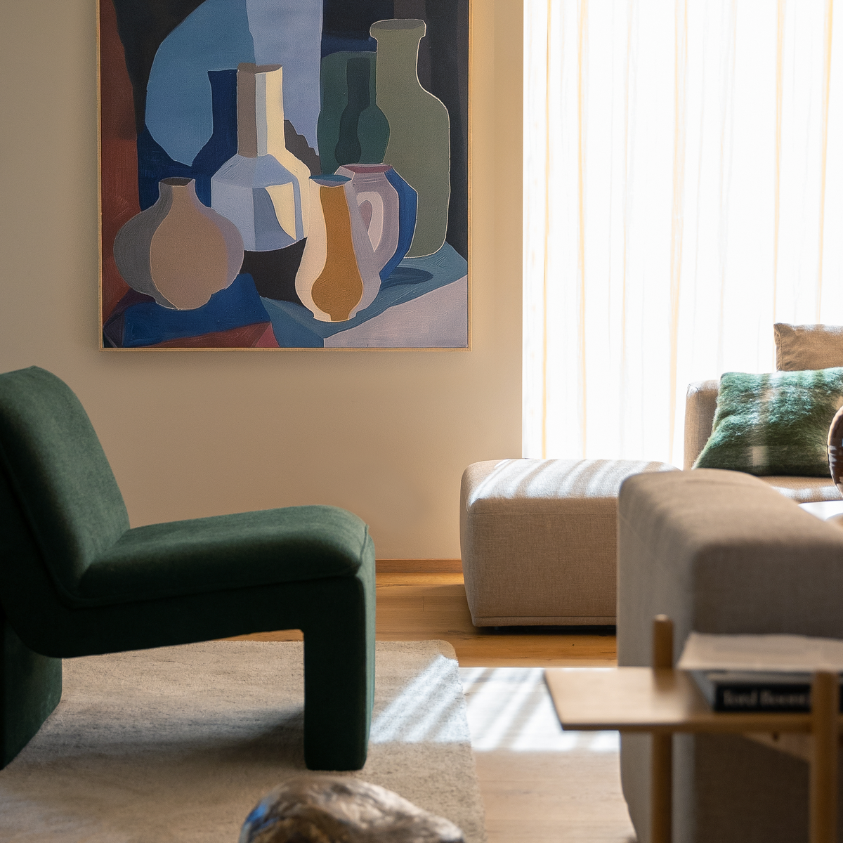 Lyst stue med grøn loungestol, beige sofa og puf, stor abstrakt vægkunst med vaser og blødt dagslys gennem lyse gardiner.