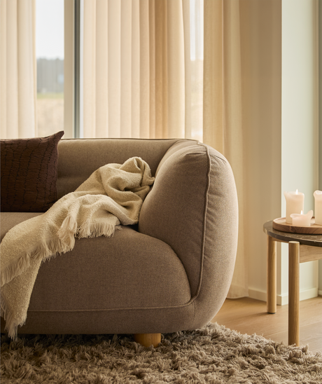 Nærbillede af et varmt, neutralt stuehjørne: brun sofa med cremefarvet plaid og pude, langluvet tæppe og lille træbord med tændte lys foran beige gardiner i dagslys.