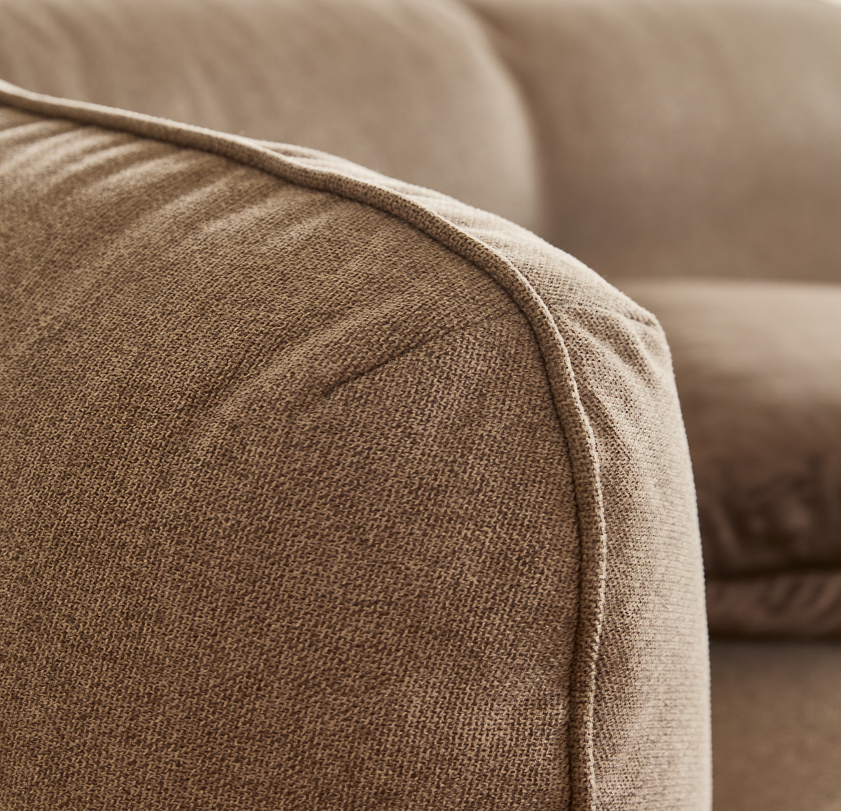 Nærbillede af beige stofsofas afrundede armlæn med synlig keder/piping.