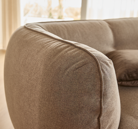 Nærbillede af rund, polstret sofaarm i beige tekstil med matchende pude i et lyst stuemiljø.