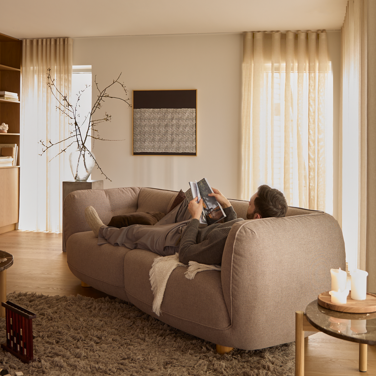 Mand ligger og læser på en beige sofa i en lys stue med lette gardiner, vægkunst, vase med grene, tæppe og lys på et sidebord.