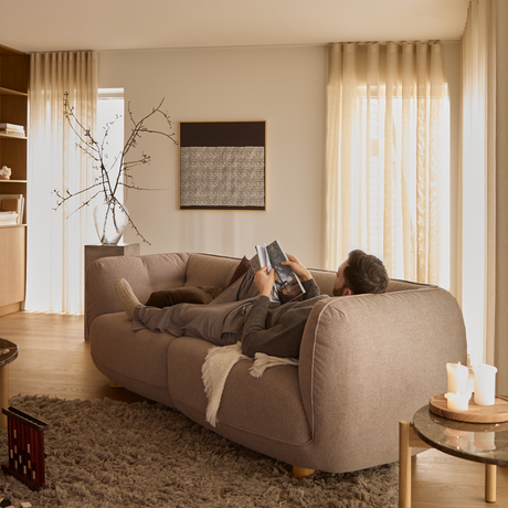 Mand ligger og læser på en beige sofa i en lys stue med lette gardiner, vægkunst, vase med grene, tæppe og lys på et sidebord.