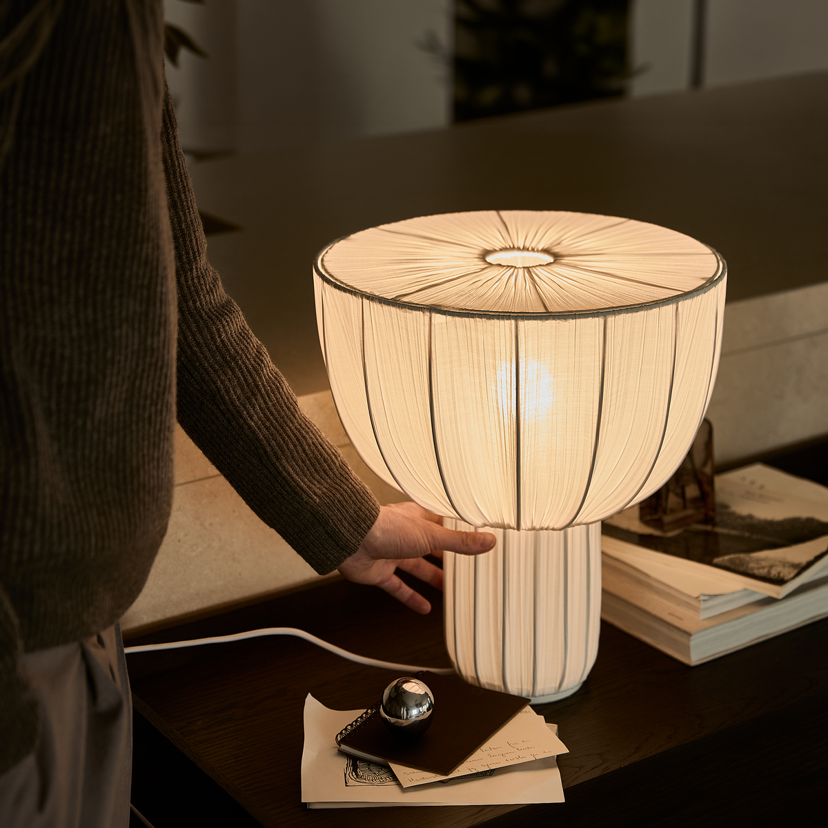 Hånd der justerer en varmglødende bordlampe med plisseret skærm på et mørkt sidebord med bøger.