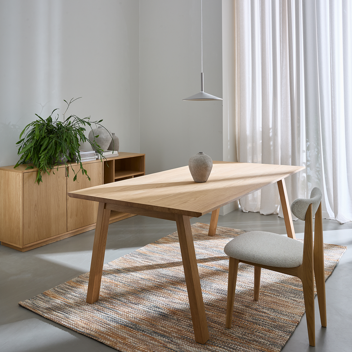 Lyst spiseområde med egetræsbord og stol på vævet tæppe, keramisk vase som midtpunkt, skænk med grøn plante og lette gardiner under en slank pendellampe.