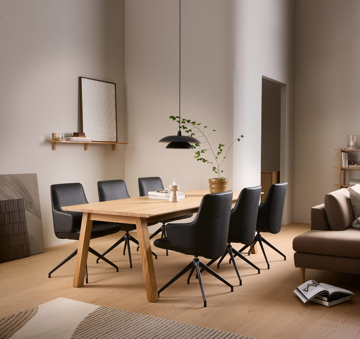 Spiseområde med rektangulært spisebord i lyst egetræ, seks sorte drejestole, sort pendellampe og en grøn plante i guldfarvet potte; bøger og lys på bordet i et lyst, roligt skandinavisk interiør.