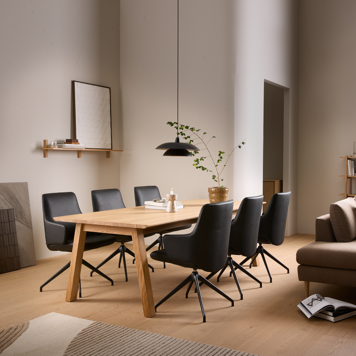 Spisestue med rektangulært bord i lys eg, seks sorte drejestole og sort pendel; bøger, lys og en grøn plante i potte på bordet i et lyst skandinavisk hjem.