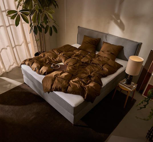 Soveværelse med grå seng, brun dyne og puder, sengebord med lampe, stor plante og varm stemningsbelysning.