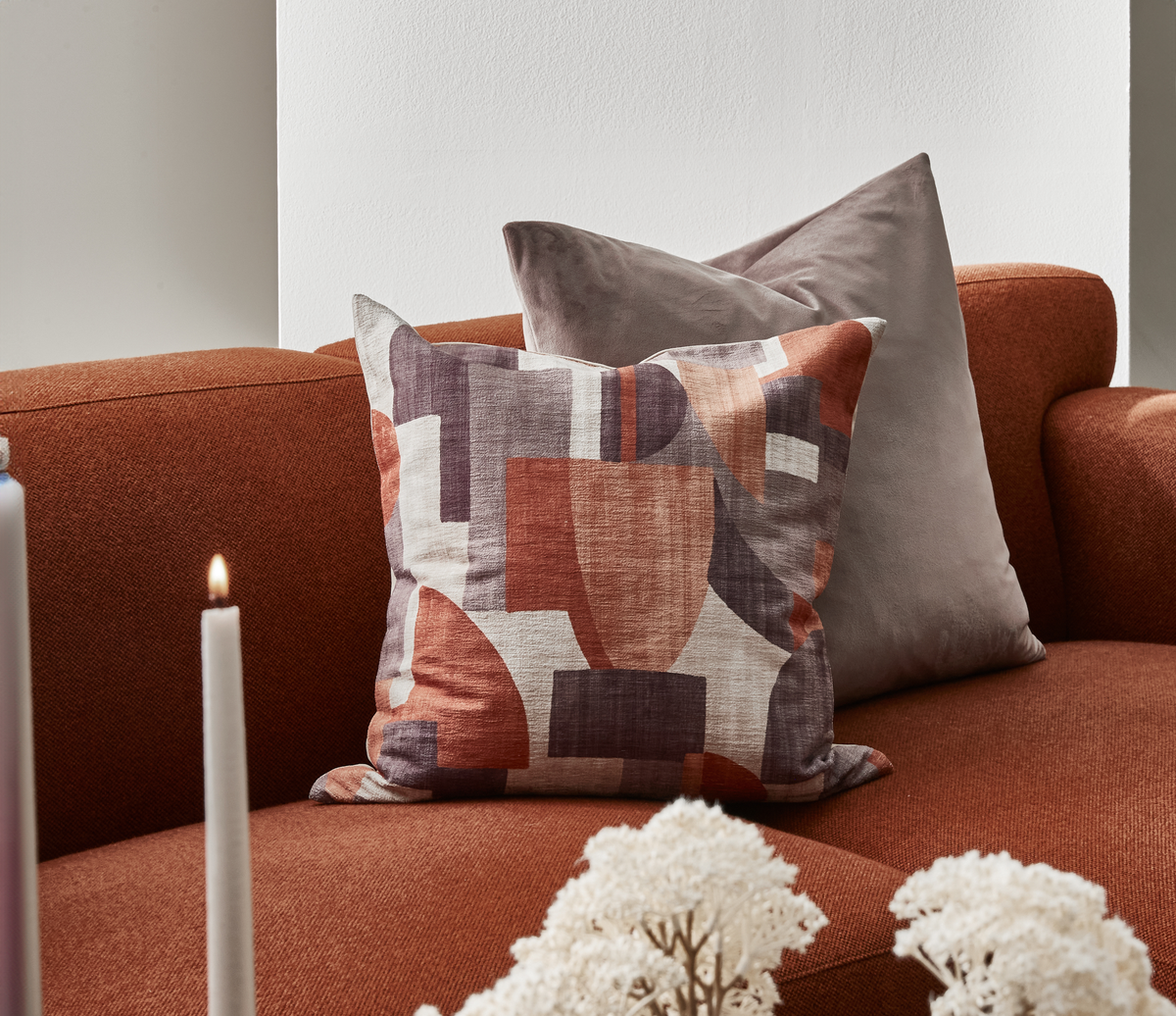 Rustfarvet sofa med to pyntepuder, den ene med abstrakt mønster i terrakotta og mørkegrå, med tændt lys og tørrede blomster i forgrunden.