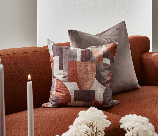 Rustfarvet sofa med to pyntepuder, den ene med abstrakt mønster i terrakotta og mørkegrå, med tændt lys og tørrede blomster i forgrunden.