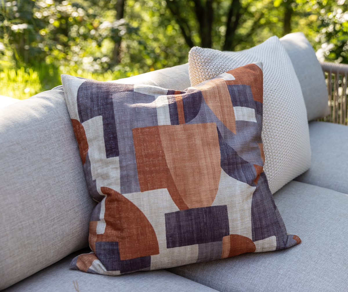 Mønstret pyntepude i rust, grå og beige på lysegrå udendørs sofa i sollys.