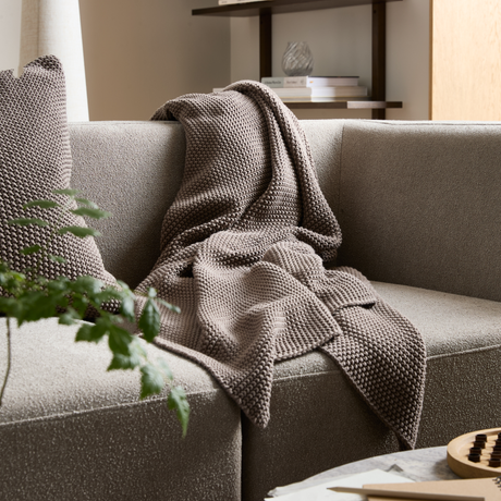 En taupefarvet strikket plaid ligger over en beige sofa med matchende pude; plante i forgrunden og bøger samt dekoration på en hylde i baggrunden.