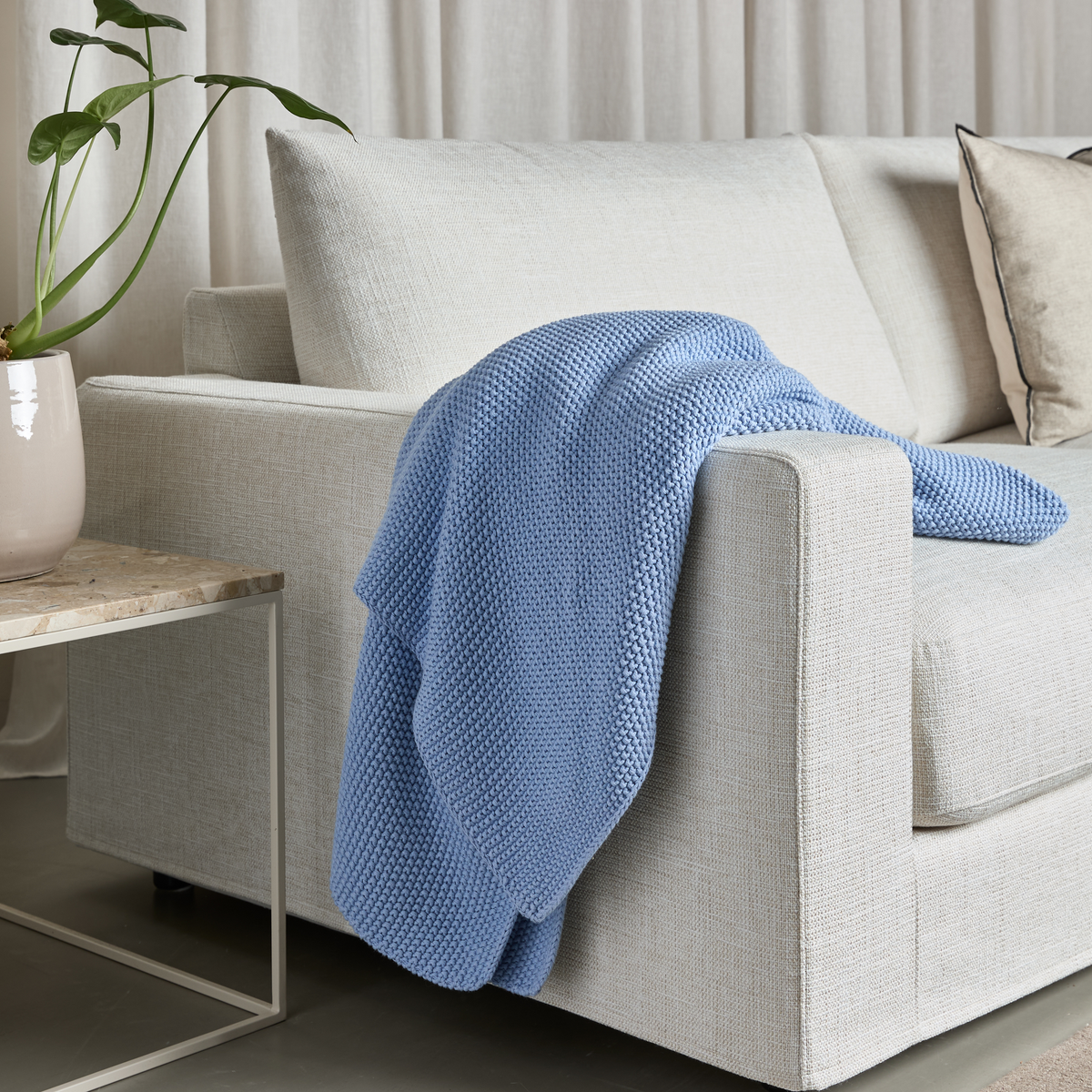 Lys beige sofa med blå strikket plaid over armlænet, marmorbord med krukke og grøn plante.