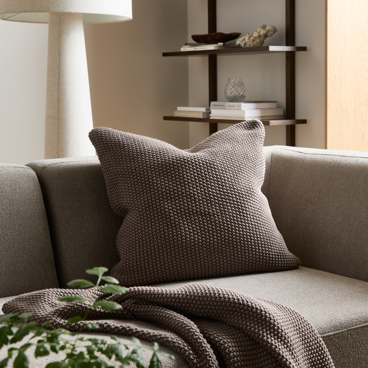 Beige sofa med grovstrikket brun pude og matchende plaid; hylde, lampe og grøn plante i en lys stue.
