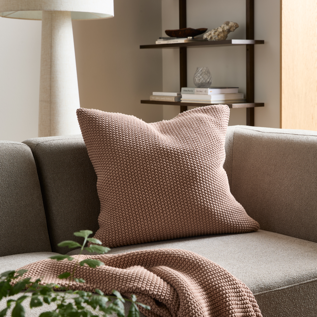 Beige sofa med en chunkystrikket pudderrosa pude og matchende plaid; gulvlampe og hylder med bøger og dekoration i en rolig stue.