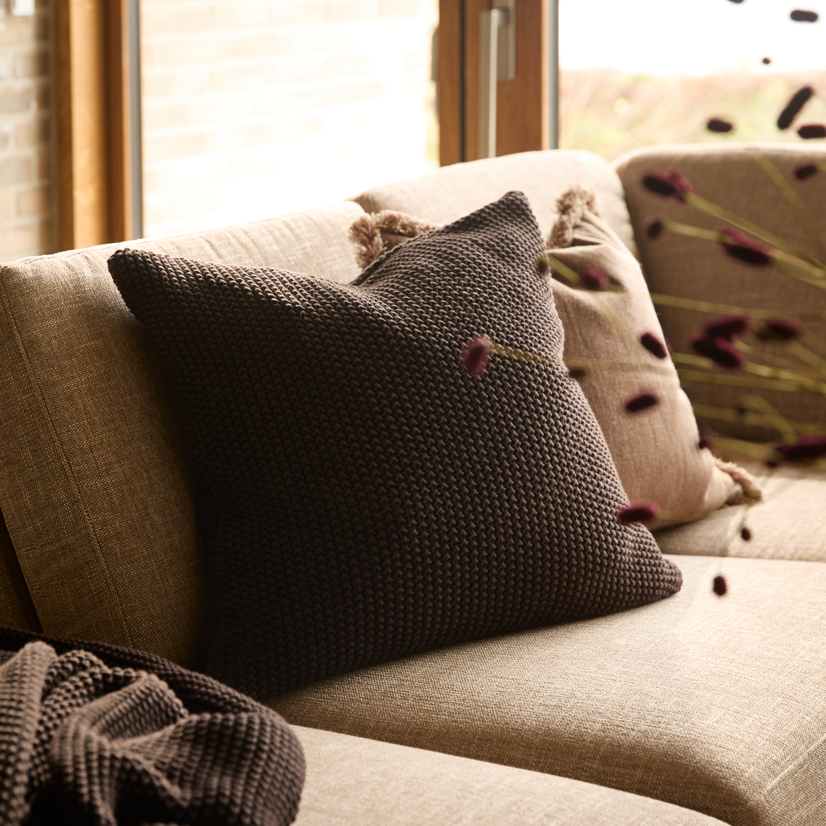 Beige sofa med en stor strikket mørkebrun pude og matchende plaid, med dagslys fra vinduet og tørrede blomster i forgrunden.