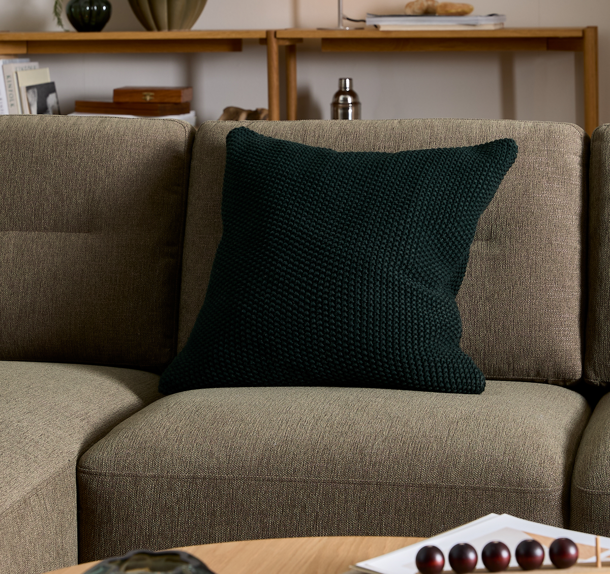 Mørkegrøn strikket pude på beige sofa i stue med træhylde og dekoration i baggrunden.
