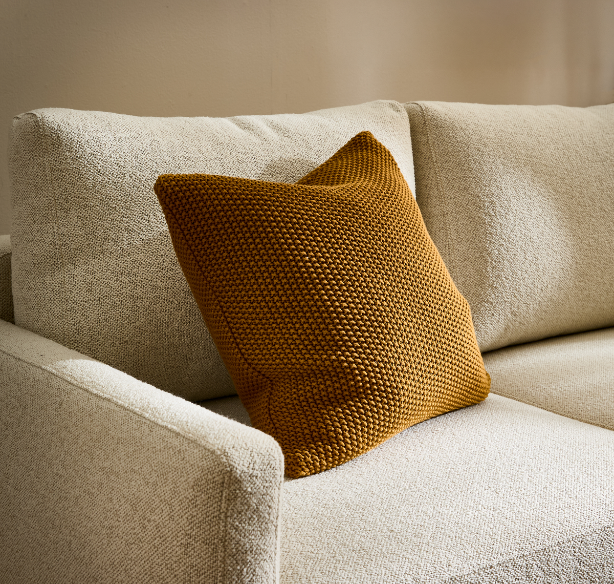 Beige sofa i tekstureret stof med okkergul strikket pude.