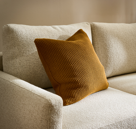 Beige sofa i tekstureret stof med okkergul strikket pude.