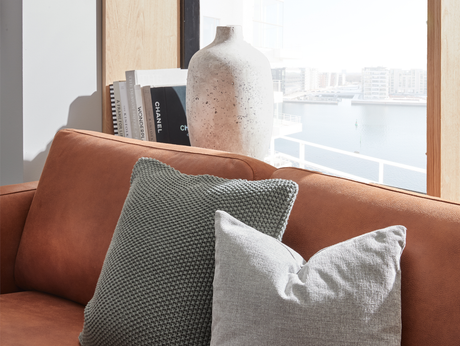 Nærbillede af en brun lædersofa med grå strik- og hørpuder, en plettet keramikvase og bøger som Chanel og Wonder ved et lyst vindue med udsigt over vand og lejligheder.
