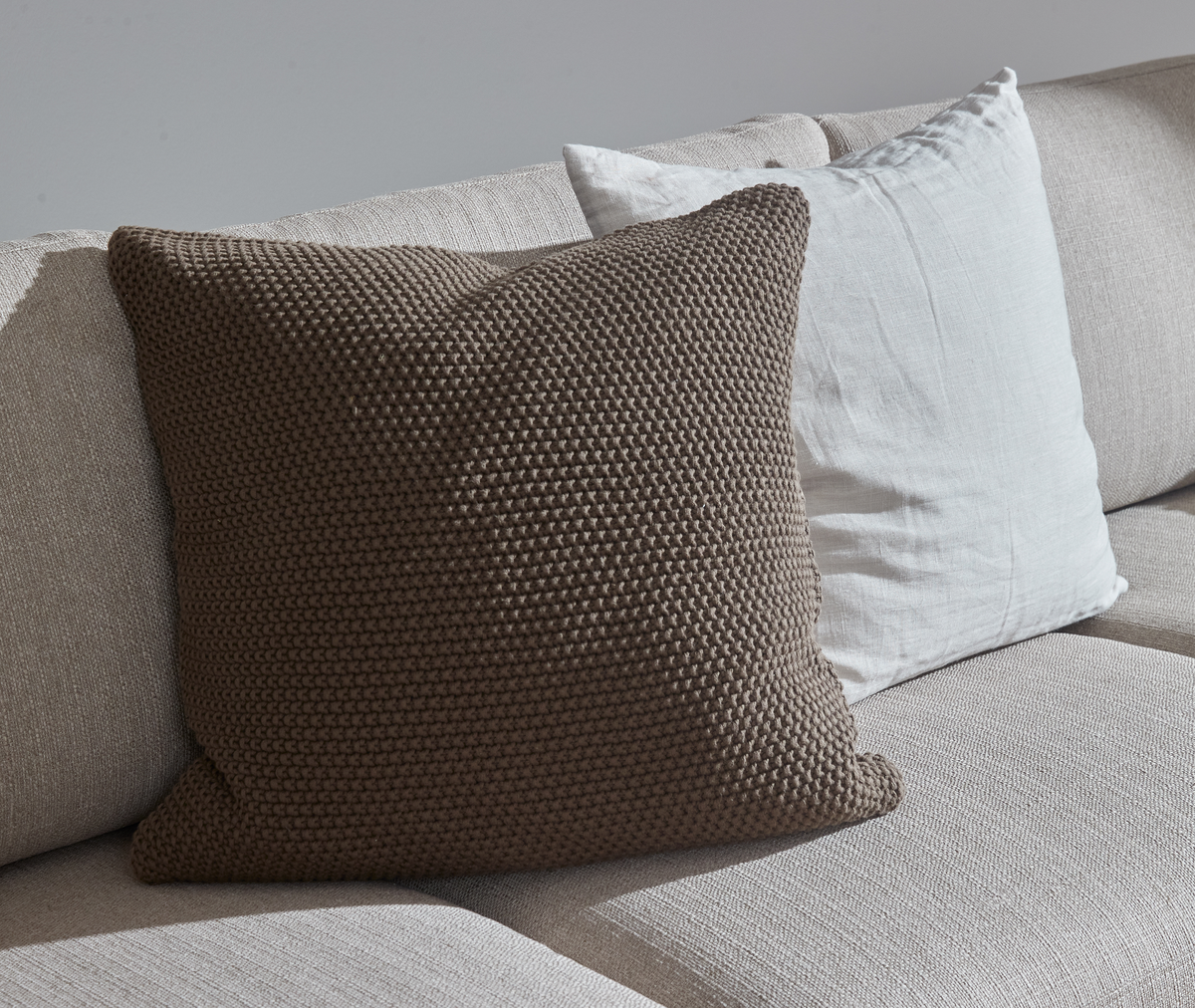 Nærbillede af beige sofa med strikket brun pude foran en hvid hørpude i blødt dagslys.