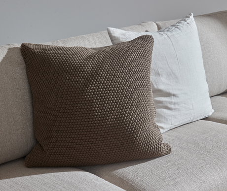 Nærbillede af beige sofa med strikket brun pude foran en hvid hørpude i blødt dagslys.