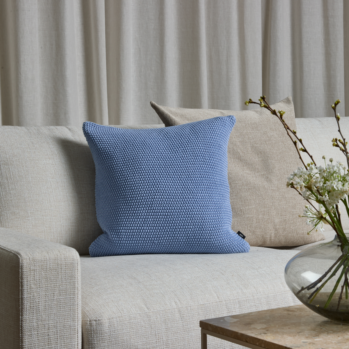 Lys beige sofa med blå tekstureret pude foran en beige pude, glasvase med hvide blomster på bord og neutrale gardiner i baggrunden.