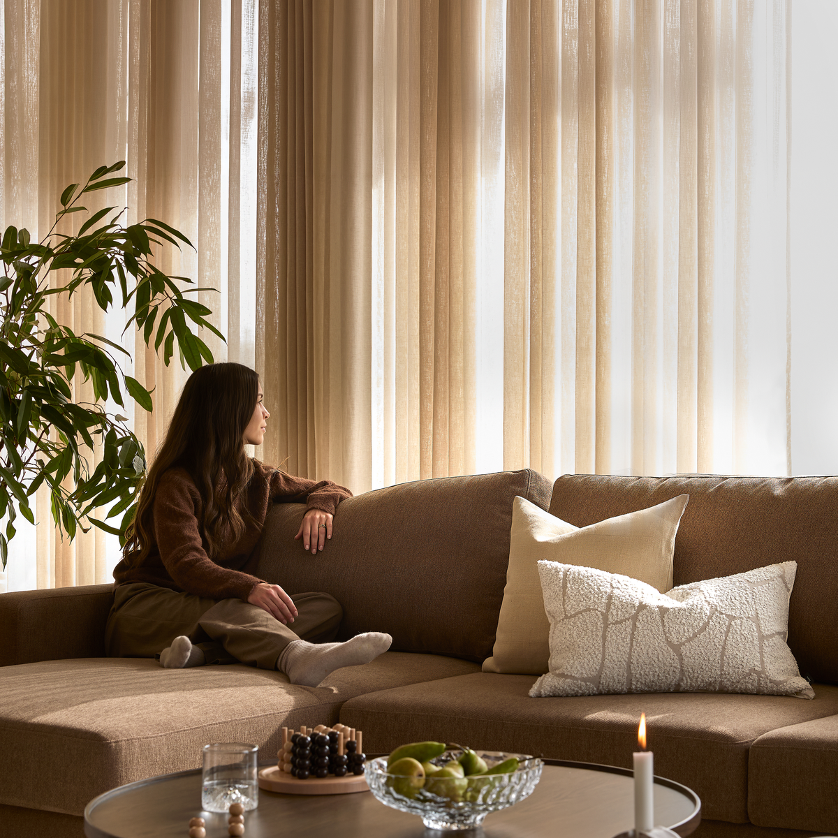 Stue med beige gardiner, brun hjørnesofa med lyse puder og et sofabord med frugtfad, glas og tændt lys; en person sidder afslappet og kigger mod vinduet.