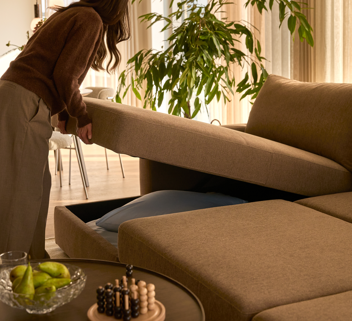 Person løfter sædet på en beige modulsofa med opbevaring til sengetøj; stue med plante og rundt sofabord med frugt og spil.