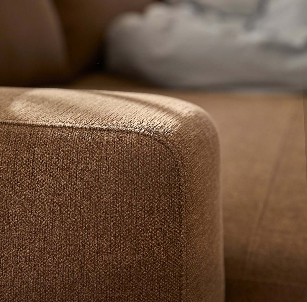 Nærbillede af brun stofsofa med afrundet armlæn i blødt dagslys.