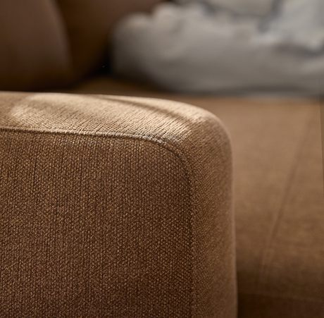 Nærbillede af brun stofsofa med afrundet armlæn i blødt dagslys.