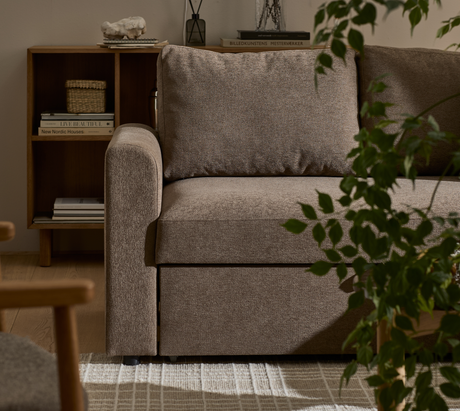 Brun stofsofa i stuen med stor pude; træreol ved siden af med bøger – Live Beautiful, New Nordic Houses, Billedkunstens Mesterværker – og lille kurv; grøn plante i forgrunden og lyst mønstret tæppe.