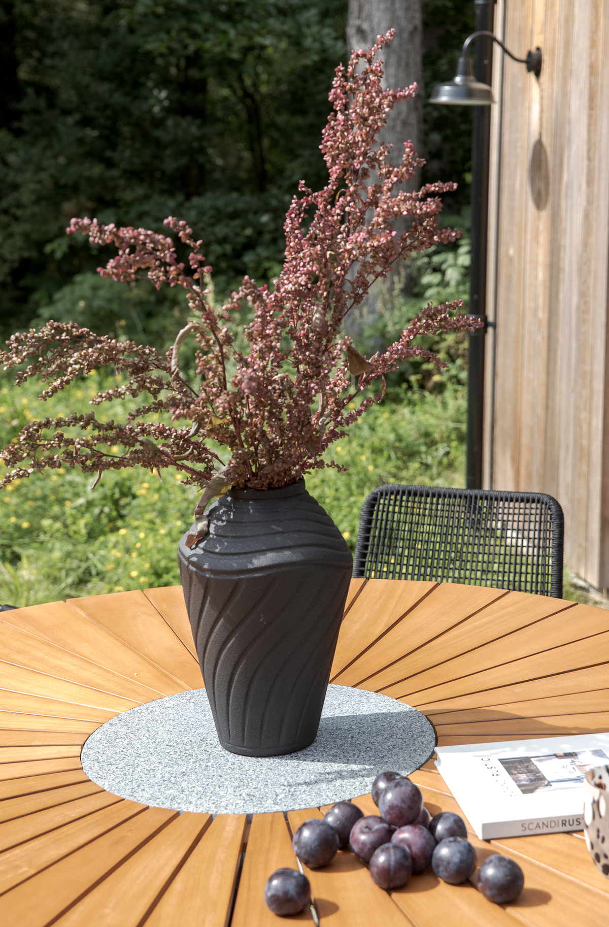 Sort, struktureret vase med tørrede grene på rundt træbord udendørs; blommer ligger ved siden af sammen med bogen Scandi Rustic.