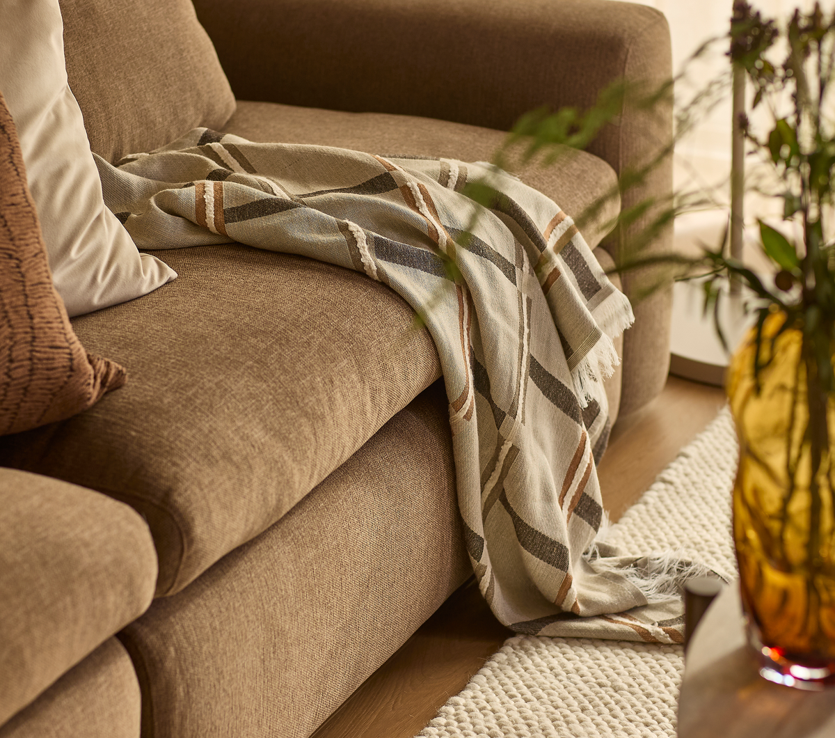 Brun stofsofa med stribet plaid over armlænet, puder og lys grovstrikket måtte; ravgul glasvase i forgrunden i en lys stue.