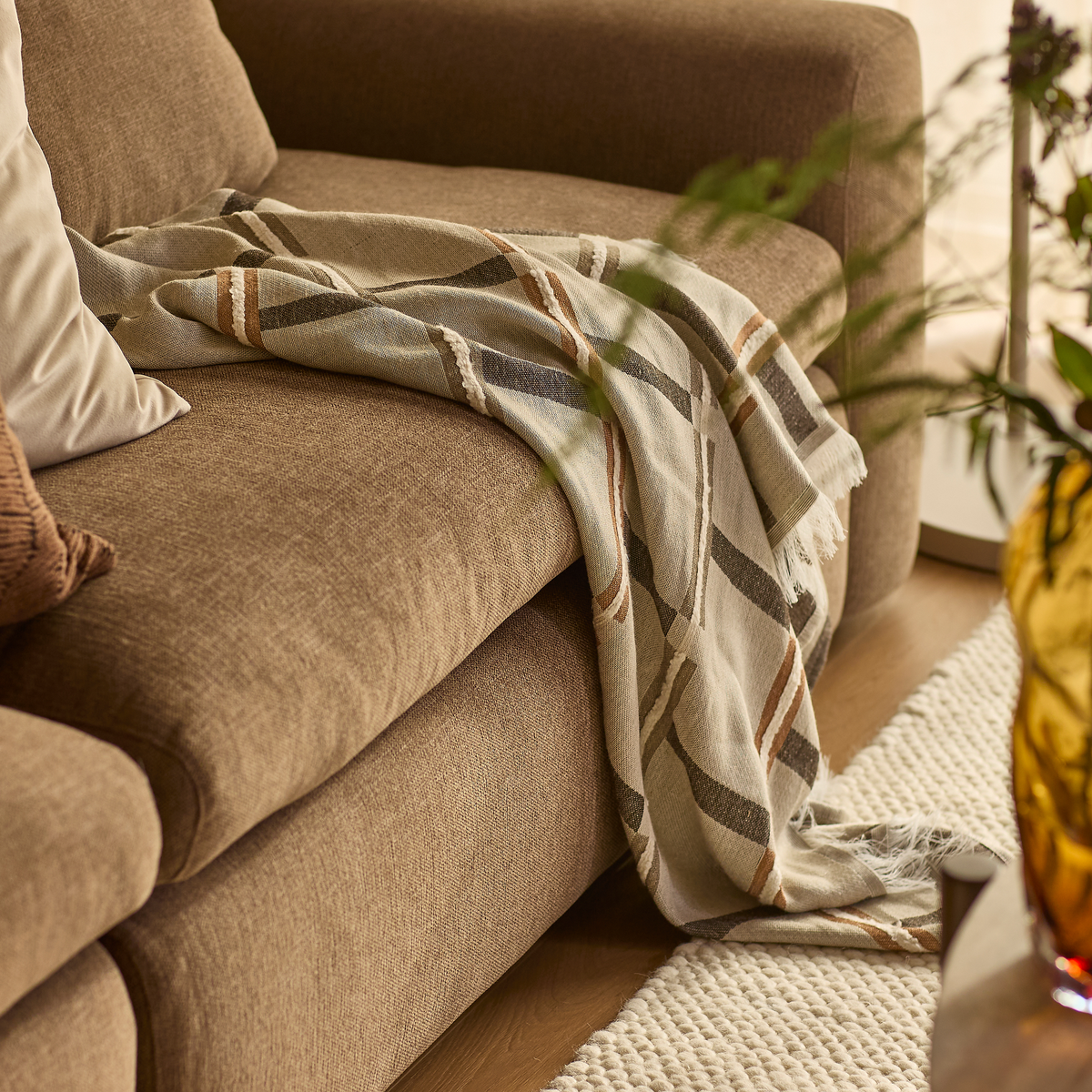Brun sofa med mønstret plaid, der hænger over kanten, pude og lys, tekstureret tæppe i en varm, solfyldt stue.