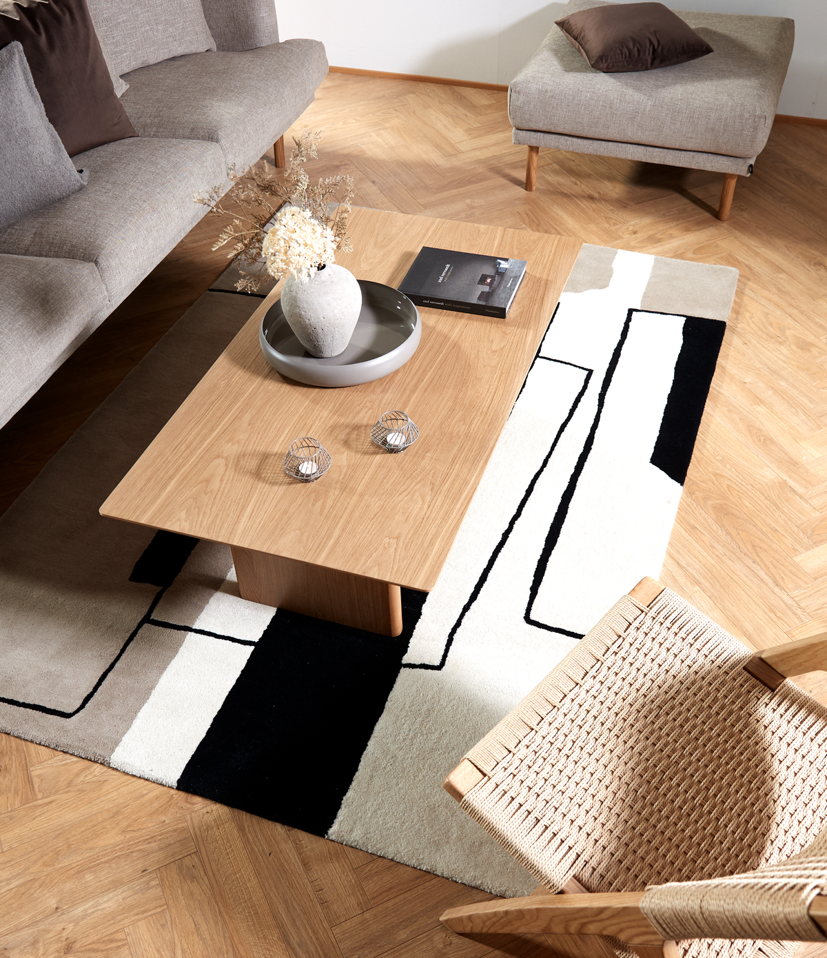 Stue med træ-sofabord på et grafisk sort-hvidt og beige tæppe, grå sofa og puf, fletstol samt vase med tørrede blomster og en bog på bordet.