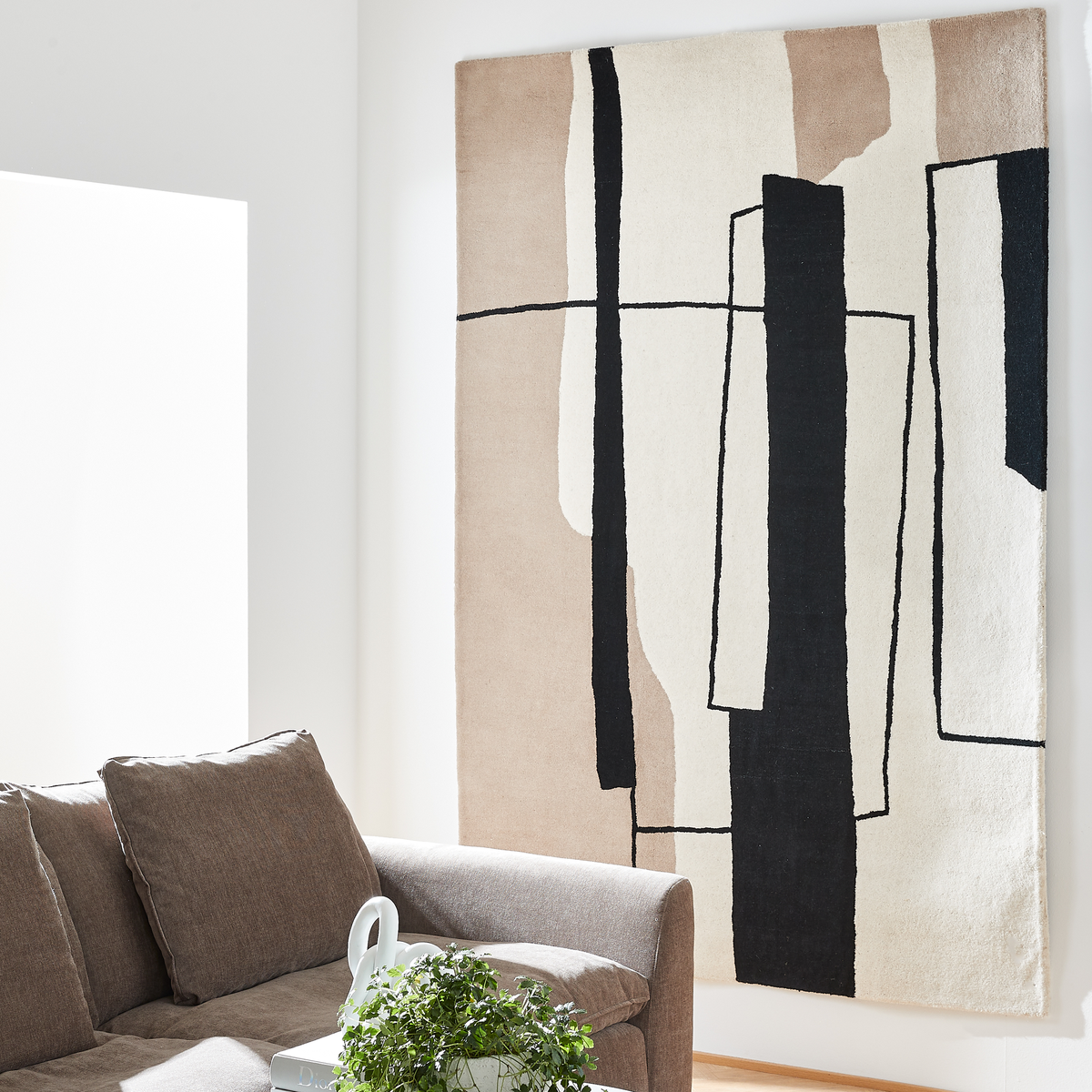 Lyst stue med brun sofa, grøn plante på bordet og et stort abstrakt vægophængt tæppe i beige, sort og hvid.