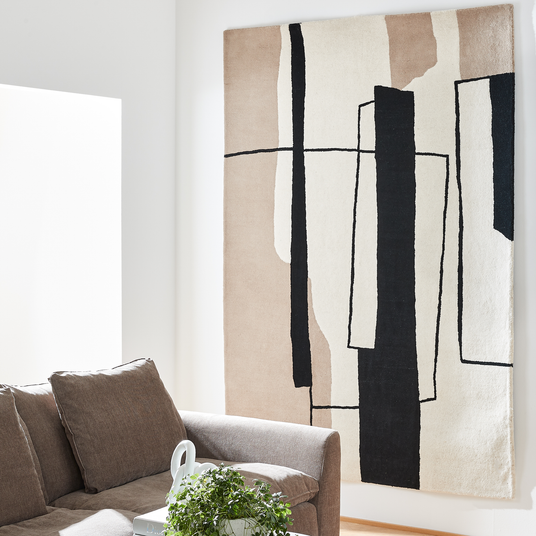 Lyst stue med brun sofa, grøn plante på bordet og et stort abstrakt vægophængt tæppe i beige, sort og hvid.
