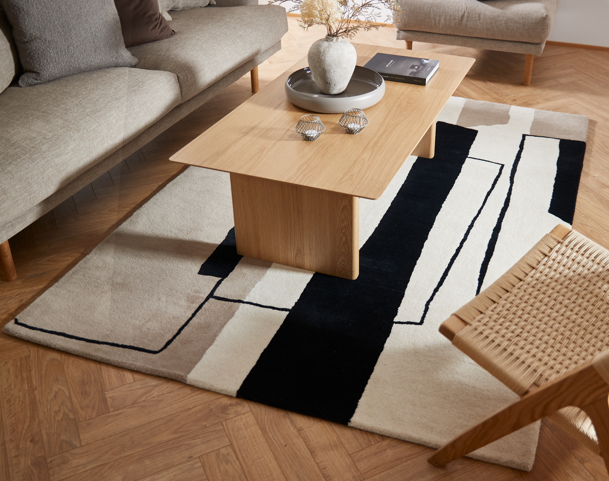 Stue med lyst sofabord i træ på grafisk sort‑beige tæppe, sofa og fletstol, vase med tørrede blomster og bog på bakke.
