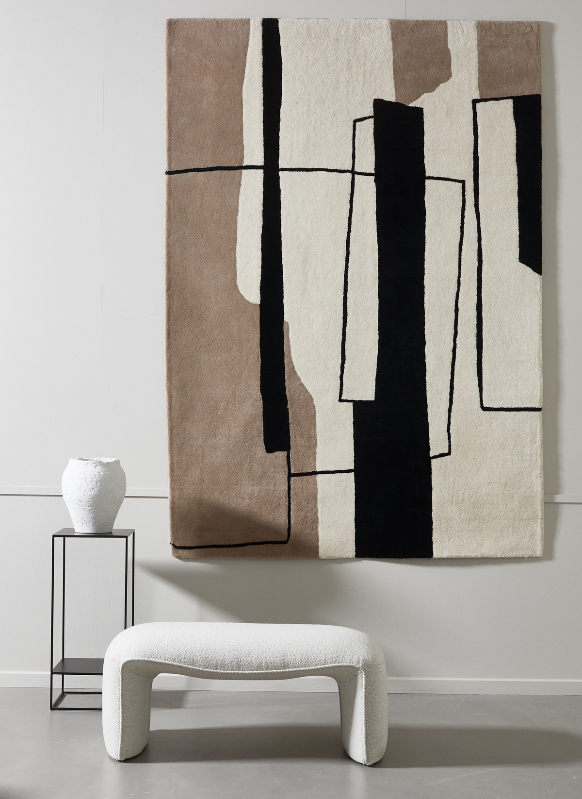 Abstrakt geometrisk tæppe i beige, creme og sort hænger på væggen over en hvid bouclébænk; ved siden af står et sort metalstativ med en hvid vase.