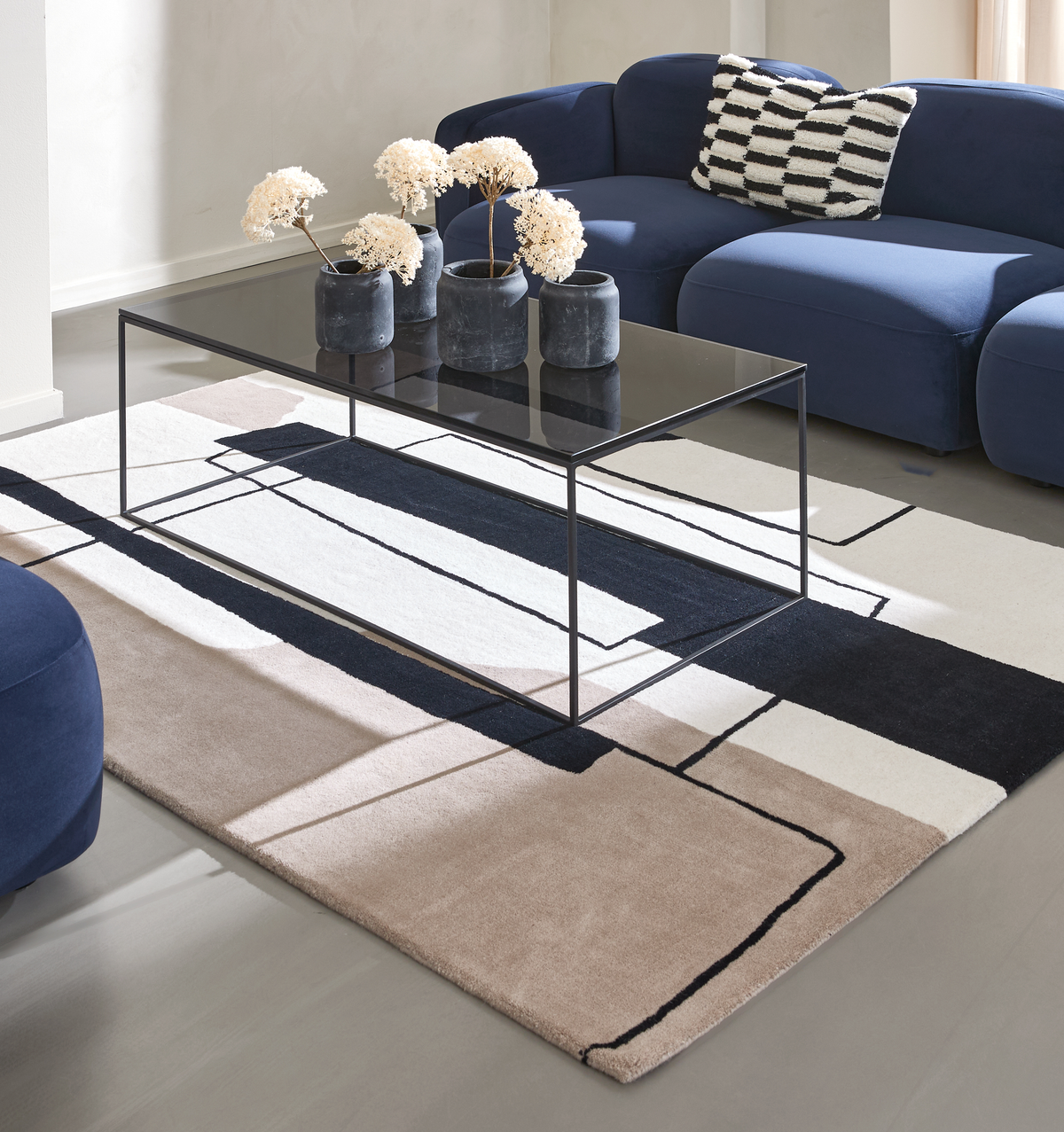 Moderne stue med mørkeblå sofa, sort glas-sofabord og grafisk tæppe i beige, sort og hvid samt vaser med tørrede blomster.