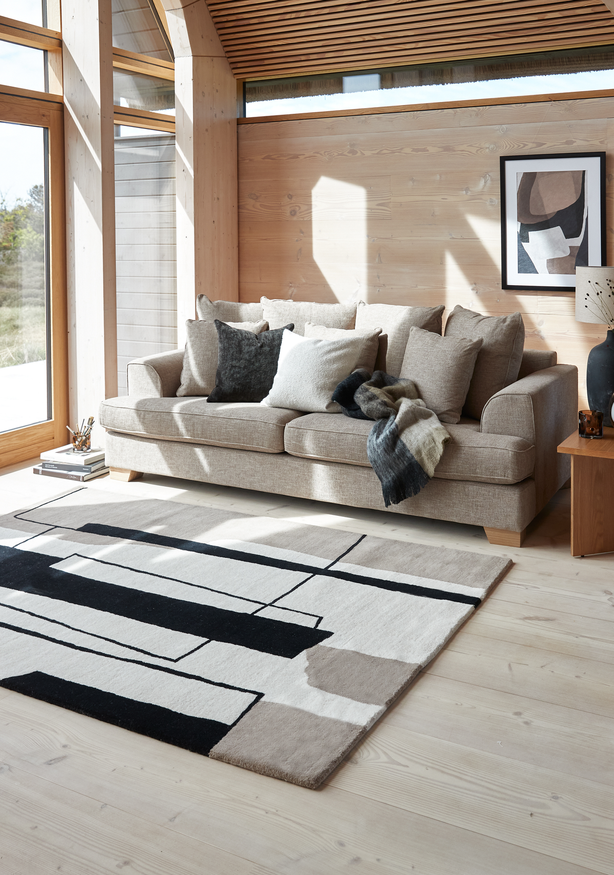 Lys skandinavisk stue med beige sofa, puder og plaid, abstrakt sort‑hvid‑beige tæppe, store trævinduer og vægkunst i sollys.
