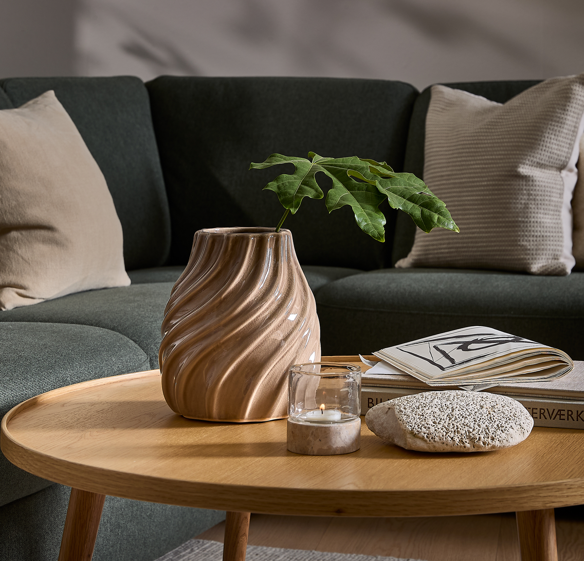 Rundt sofabord i træ med bølget beige vase med grøn gren, tændt fyrfadslys, bøger og en lys sten foran en mørkegrøn sofa med neutrale puder.
