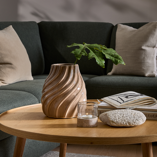 Lyst sofabord i træ med bølget keramikvase og grøn kvist, tændt fyrfadslys i glas, dekorativ sten samt åben blad på en bog med ryggen “BILDSERVER”, foran mørkegrøn sofa med beige og lyse puder.
