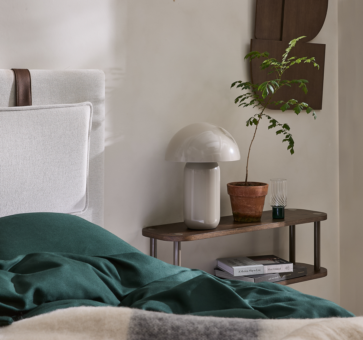 Sengebord med svampeformet bordlampe, terracottakrukke med lille plante og bøger; seng med grønt sengetøj i forgrunden.