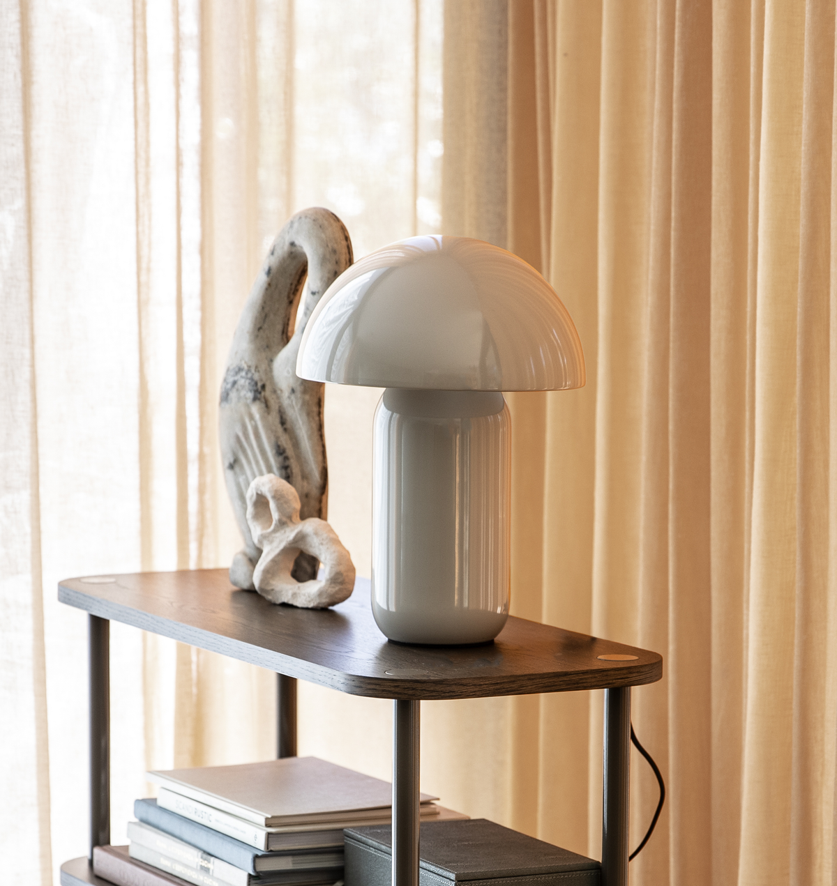 Hvid svampeformet bordlampe på hylde med skulpturel dekoration og bøger foran beige gardiner.
