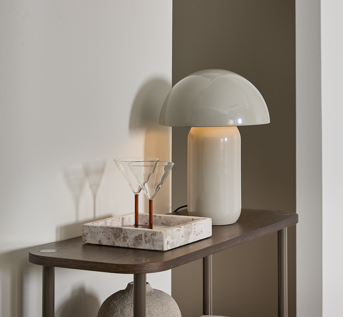 Blank hvid bordlampe med kuppelformet skærm og varm glød ved siden af to martiniglas i en terrazzo-bakke på et mørkt konsolbord mod lyse vægge.