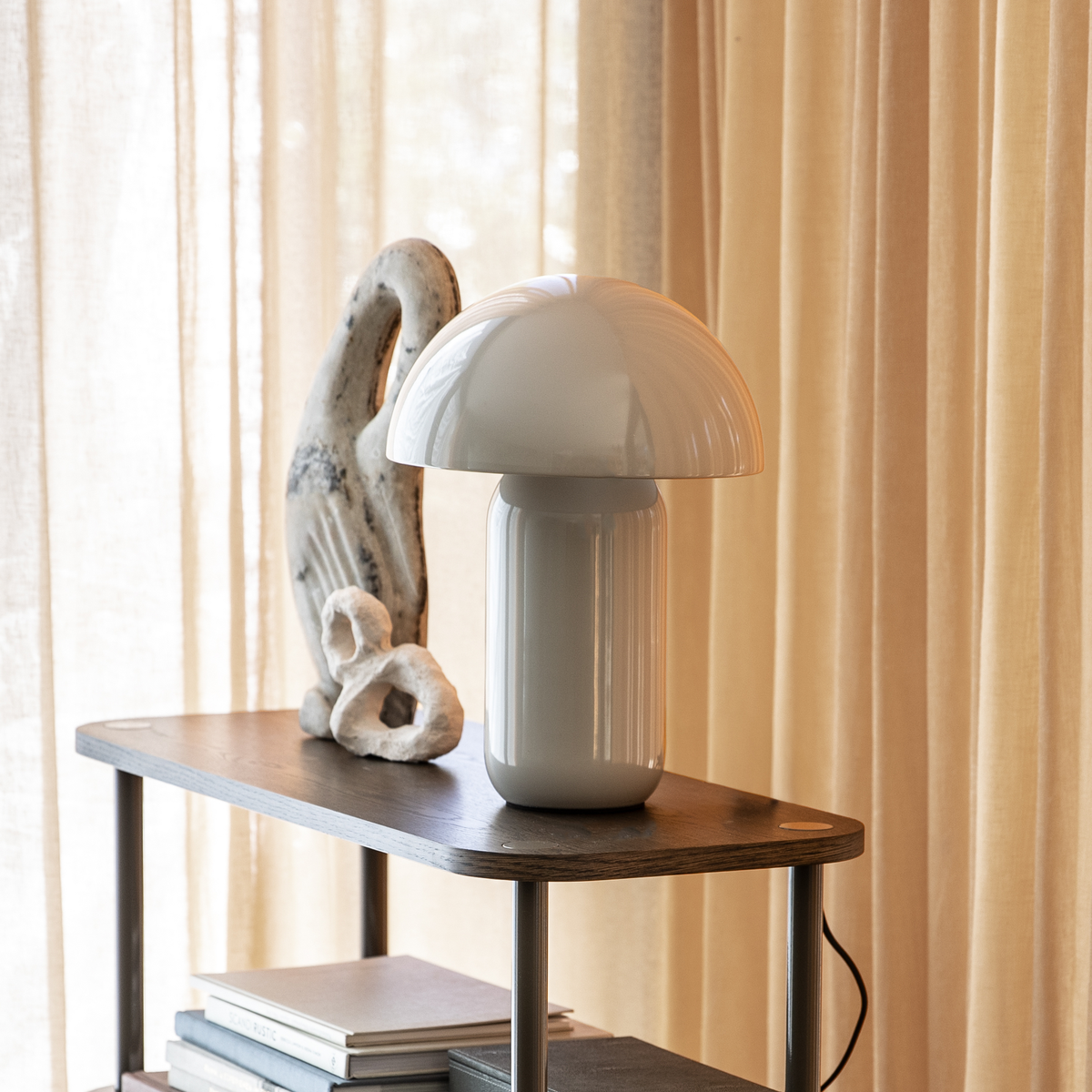 Hvid svampeformet bordlampe på en træhylde med skulptur og bøger foran beige gardiner.