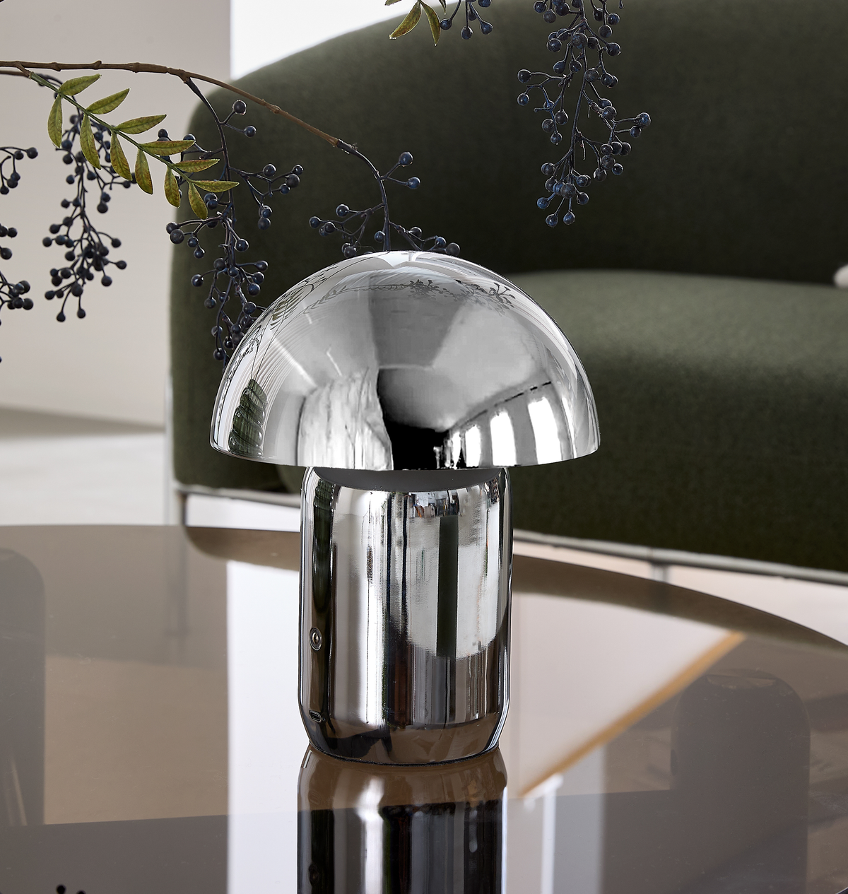 Forkromet svampeformet bordlampe på et glasbord med grøn sofa og bærgrene i baggrunden.